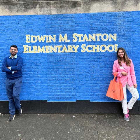 Two individuals standing in front of a blue brick wall with the text “Edwin M. Stanton Elementary School” painted in large yellow letters.