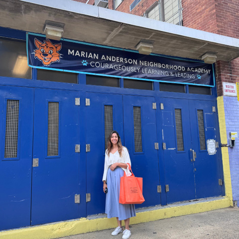 A person stands in front of a blue double door entrance to the Marian Anderson Neighborhood Academy, which has a sign featuring an orange fox mascot and the words “Courageously Learning & Leading