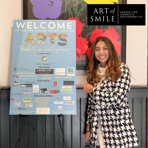 A person stands next to a sponsorship board for the 11th Annual Spring into the Arts event, featuring the Art of Smile - Centre for Cosmetic Orthodontics as a key sponsor among various logos of local businesses in Philadelphia.